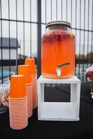 A beverage dispenser that is filled with a bright orange liquid stands next to several stacks of orange cups, ready to be used for serving delicious drinks at gatherings and events photo