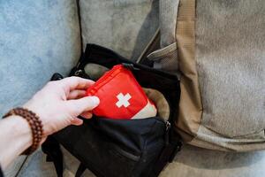 A person takes a small first-aid kit with medicines with him on the road, puts a red first-aid kit in the pocket of his bag. photo