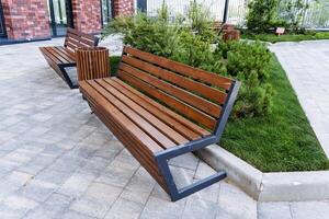 In a park, there is a line of wooden benches with a brick building in the background. The setting includes green grass and a wooden structure that complements the outdoor scenery photo