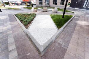 In the middle of the park, there is a triangular feature surrounded by greenery and urban design elements like buildings and roads. The park also includes plants and grass photo