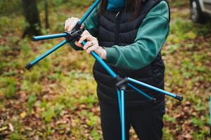 A confident woman stands with pride in a serene wooded area, firmly holding a blue tripod, fully prepared for an adventure amidst the lush greenery and towering majestic trees photo