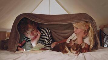 Brother is reading a book using his phone light under a blanket with his sister and pet chihuahua video