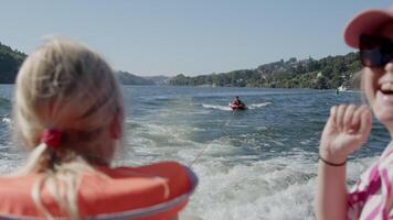 Blonde woman with her daughter in a life jacket, filmed from the back, watching another person tubing on a lake with forested hills in the background video