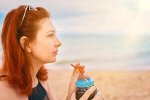 Red-haired girl in a swimsuit is sitting on the beach with a cocktail and look away. Calm woman on vacation. Holidays at the sea. Vintage sunglasses on the head of a having a rest lady. photo