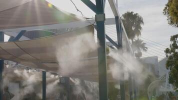 Water vapor is being sprayed from a series of nozzles on a metal structure with a fabric roof, in an effort to keep tourists cool video