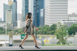 A young professional woman walking confidently in an urban setting with a city skyline and park in the background, holding a tablet, dressed in business casual attire, showing determination photo
