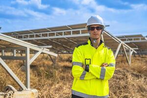 A confident solar energy technician, wearing a helmet and reflective jacket, stands with arms crossed in front of an array of photovoltaic panels, under a bright and clear blue sky. photo