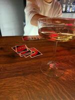 A stylish cocktail glass sits on a wooden bar, with colorful UNO cards in the foreground. A hand reaches out, enhancing the playful ambiance of the evening. photo