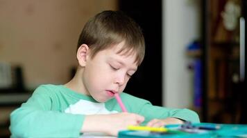 cute and smiling caucasian boy draws with colored pencils while sitting at the table. the boy paints the drawing with a pink pencil. Drawing lessons. Art therapy for stress relief. video