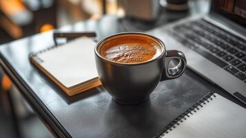 A coffee cup with coffee stands on a black leather desk next to a notebook and a notepad photo