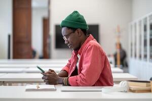 Smiling African guy student using smartphone during study break, sitting at desk in library photo