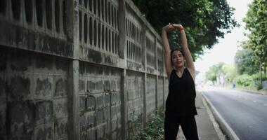 Handheld shot, Young Asian woman in sportswear stretching to warm up before exercising outdoors on the sidewalk next to a wall in the city. Health and Wellbeing video