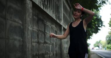 Handheld low angle view shot, Young Asian woman wear wireless headphones leaning against wall and wiping her sweat off after a workout on a city sidewalk. Health and Wellbeing. video
