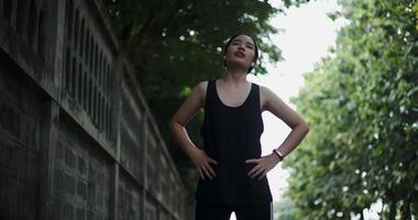 Handheld low angle view shot, Young woman in sportswear and wireless headphones listening to music while stretching to warm up before an outdoor workout on a sidewalk next to a wall video