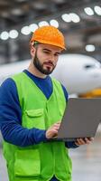 Aircraft Maintenance Technician Working on Laptop in Hangar with Airplane Background, Safety Gear and High-Visibility Vest, Technology and Aviation Industry photo