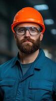 Bearded man wearing safety glasses and orange hard hat in a blue uniform. Industrial setting with blurred background. Portrait photography for workplace safety and construction concept photo