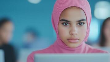 Nonbinary Teenager in Pink Hijab Focused on Laptop Screen in Classroom Setting with Blue Background Creating a Thoughtful and Contemplative Atmosphere for Educational and Learning Concepts photo