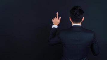 Man in a suit pointing upward with one finger, standing against a dark background. Rear view showing professional attire and gesture indicating direction or emphasis. photo