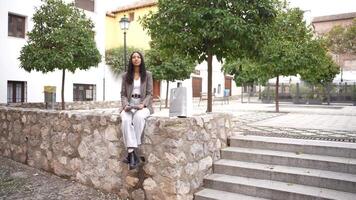 A contemplative woman sits on a stone wall in a scenic town square, reflecting on life video
