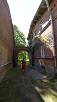A woman in a red dress walks through an overgrown courtyard between two brick buildings of an old house, heading toward a rustic archway. The lush greenery creates a peaceful and tranquil atmosphere. video