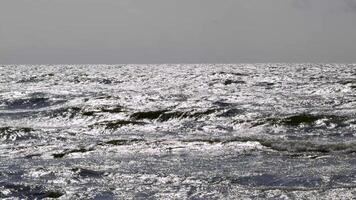 A high-angle view of the open sea with choppy waves sparkling in bright sunlight. The reflective water contrasts with the overcast grey sky, showcasing the power and motion of the ocean video