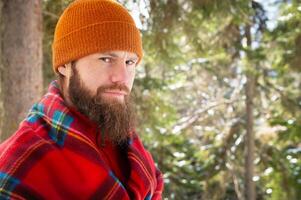 Bearded man smiling while standing among lush green forest with happy expression in checkered red blanket photo