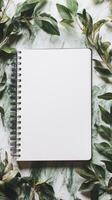 A blank notebook sits on a table with green leaves surrounding it photo