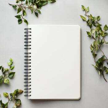 A spiral bound notebook with a white cover sits on a table with green leaves photo