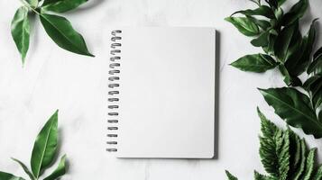 A spiral notebook sits on a white background with green leaves surrounding it photo