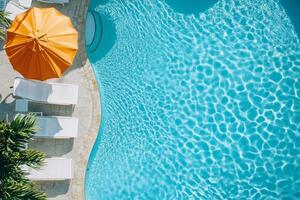A pool with a large umbrella and several lounge chairs. The water is calm and the sun is shining brightly photo
