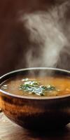 A steaming bowl of soup garnished with fresh herbs sits on a rustic wooden table. The warm, inviting aroma and vibrant colors depict comfort food at its finest. photo