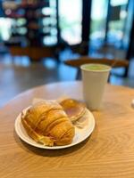 Cozy breakfast scene with a croissant, pancake and matcha latte served on a wooden table. Blurred background of a cafe adds depth and evokes a sense of warmth and relaxation in a casual dining setting photo