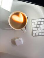 Cup of coffee with a heart-shaped reflection for Valentines day, placed on a clean white desk. Soft lighting and minimalistic workspace. Love and technologies concept. photo