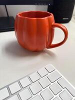 Cozy autumn workspace with a vibrant pumpkin shaped mug on a clean desk. Minimalist decor and warm tones create a relaxing atmosphere, perfect for work or leisure during the fall season photo