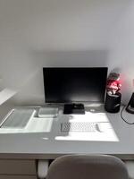 Minimalist white workspace with a computer monitor, keyboard, and other tech accessories on a clean desk. Bright sunlight streaming through the window highlights the neat and organized setting photo
