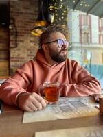 Bearded man in a peach hoodie enjoys a glass of tea in a cozy cafe. Warm string lights and rustic decor, creating a relaxed and intimate mood in restaurant photo