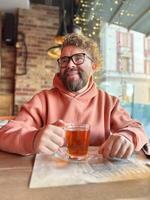 Bearded man in a peach hoodie enjoys a glass of tea in a cozy cafe. Warm string lights and rustic decor, creating a relaxed and intimate mood in restaurant photo