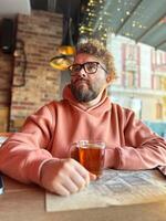 Cheerful man in a peach hoodie smiles warmly while holding a glass of tea. Cozy cafe atmosphere is accentuated by warm string lights and rustic wooden decor, creating a relaxed and inviting vibe in photo