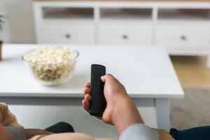 Hand holds a remote control while a bowl of popcorn sits on a white table in a modern living room. Relaxing and cozy atmosphere, perfect for watching movies or streaming content at home photo