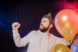 Joyful birthday man wears paper cone hat dancing and sing and stands near colorful helium balloons enjoys party and expresses happiness - holidays and winner concept photo