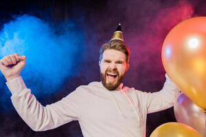 Joyful birthday man wears paper cone hat dancing and sing and stands near colorful helium balloons enjoys party and expresses happiness - holidays and winner concept photo