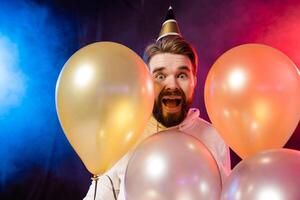 Joyful birthday man wears paper cone hat dancing and sing and stands near colorful helium balloons enjoys party and expresses happiness - holidays and winner concept photo