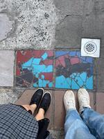 Striking mosaic of bold red and blue patterns stands out on a concrete sidewalk. Two pairs of shoes frame the image, emphasizing human interaction with the artistic cityscape photo