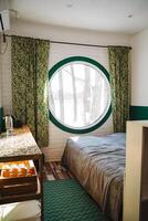 The image depicts a cozy bedroom featuring a comfortable bed and a charming round window that adds a unique touch to the overall decor. It has elements of wood and textile photo