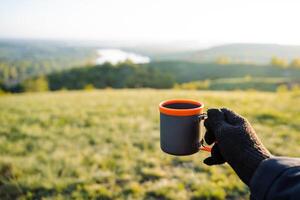 A person is standing in a grassy field, holding a cup of coffee with a joyful expression. The sky is clear, and plants surround the happy individual in the natural environment photo