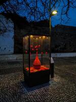 Dramatic nighttime shot of a glass display case showcasing a vibrant red ceramic vase and smaller hanging ornaments. The cobblestone setting and ambient lighting create a captivating, artistic mood. photo