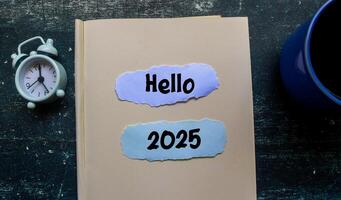 Hello 2025 text on notebook with alarm clock on wooden table. Welcoming the new year 2025 photo