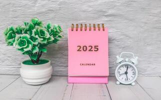 Pink calendar 2025 standing on office desk with flower pot and alarm clock. New year 2025 with office desk concept with grey background. photo