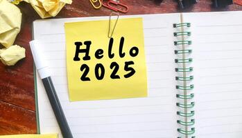 Yellow sticky note with Hello 2025 written on a notebook with stationery. Welcoming the new year 2025 concept photo