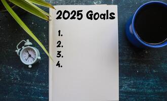 A person writes 2025 Goals in a notebook surrounded by coffee and a clock. Planning to do list for 2025, life goals for 2025 photo
